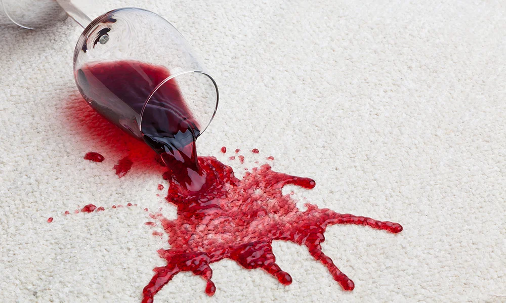 Tipped wine glass spilling red wine onto light carpet.